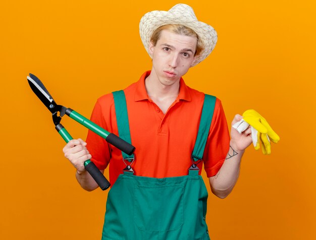 Hombre joven jardinero vestido con mono y sombrero sosteniendo cortasetos y guantes de goma