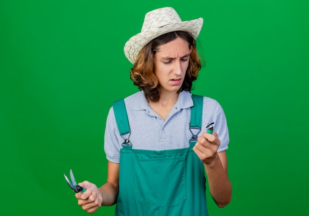 Hombre joven jardinero vestido con mono y sombrero sosteniendo equipos de jardinería