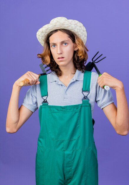 Hombre joven jardinero vestido con mono y sombrero sosteniendo mini rastrillo y azadón