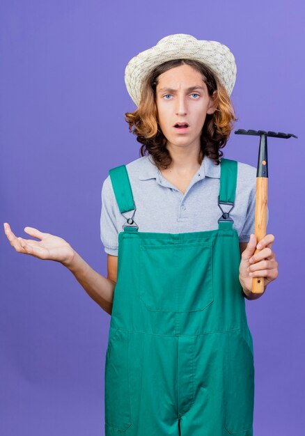 Hombre joven jardinero vestido con mono y sombrero sosteniendo mini rastrillo con brazo