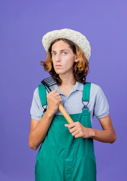 Hombre joven jardinero vestido con mono y sombrero sosteniendo mini rastrillo con cara seria