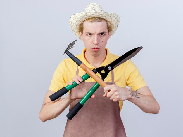 Foto gratuita hombre joven jardinero vestido con mono y sombrero sosteniendo mini rastrillo y cortasetos cruzando las manos con cara seria