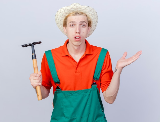 Hombre joven jardinero vestido con mono y sombrero sosteniendo mini rastrillo mirando a la cámara encogiéndose de hombros confundido de pie sobre fondo blanco.