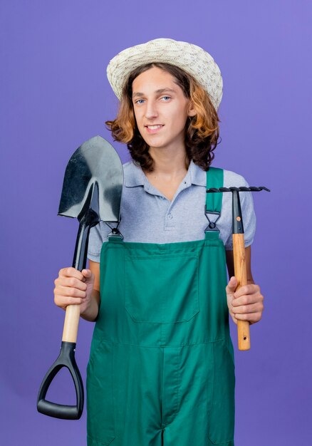 Hombre joven jardinero vestido con mono y sombrero sosteniendo mini rastrillo y pala