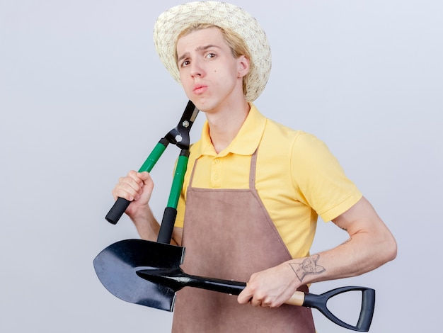 Foto gratuita hombre joven jardinero vestido con mono y sombrero sosteniendo pala y cortasetos con cara seria