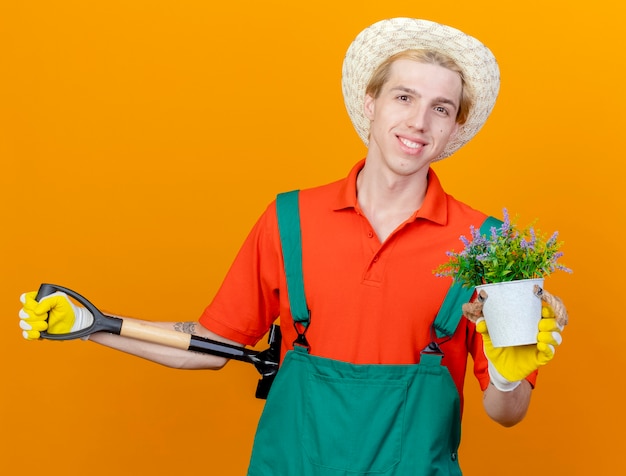 Hombre joven jardinero vestido con mono y sombrero sosteniendo pala mostrando en maceta