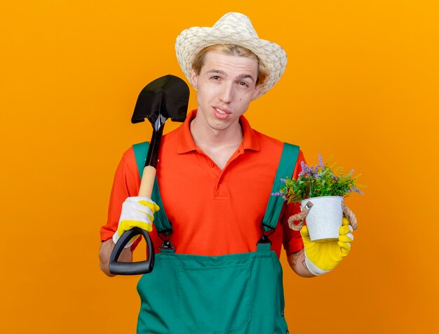 Hombre joven jardinero vestido con mono y sombrero sosteniendo pala y planta en maceta