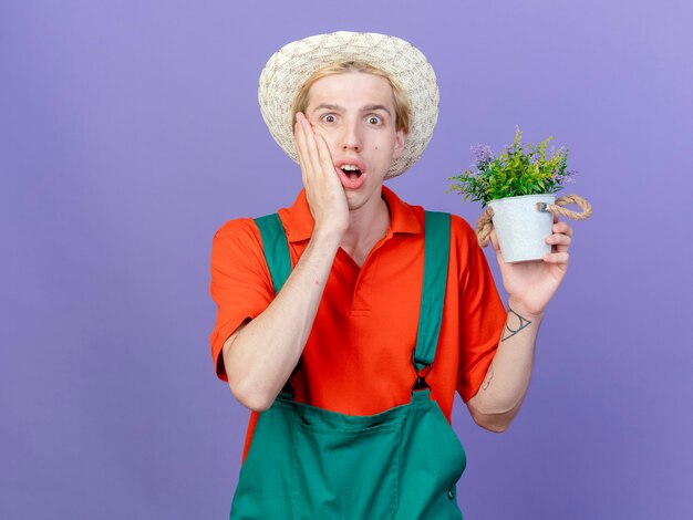 Hombre joven jardinero vestido con mono y sombrero sosteniendo planta en maceta está sorprendido