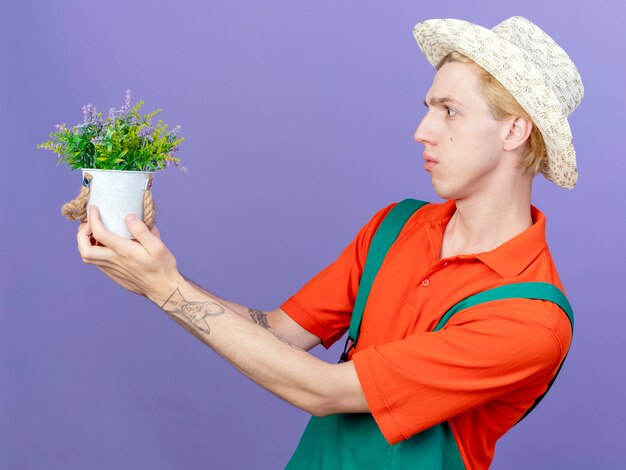 Hombre joven jardinero vestido con mono y sombrero sosteniendo planta en maceta mirando confundido