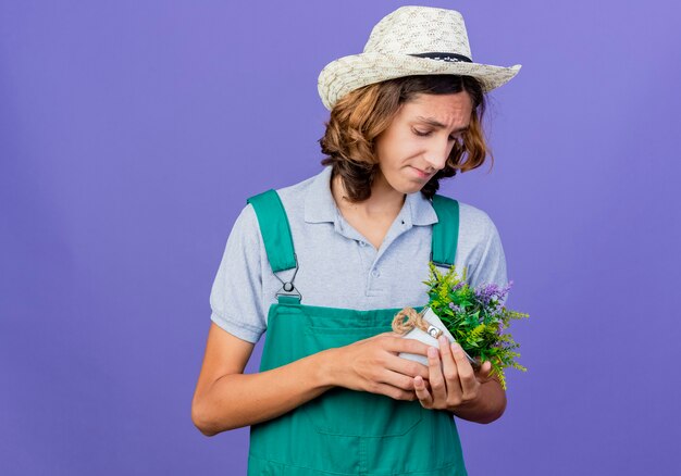 Hombre joven jardinero vestido con mono y sombrero sosteniendo planta en maceta