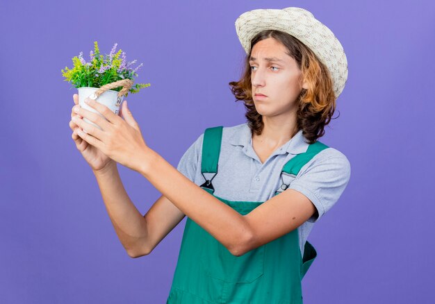 Hombre joven jardinero vestido con mono y sombrero sosteniendo planta en maceta