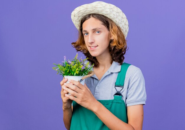 Hombre joven jardinero vestido con mono y sombrero sosteniendo planta en maceta