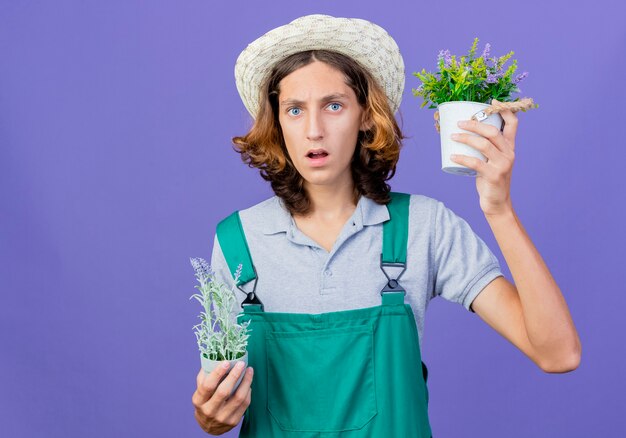 Hombre joven jardinero vestido con mono y sombrero sosteniendo plantas en macetas confundidas