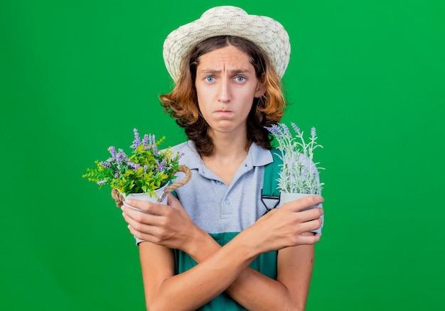 Hombre joven jardinero vestido con mono y sombrero sosteniendo plantas en macetas con expresión triste