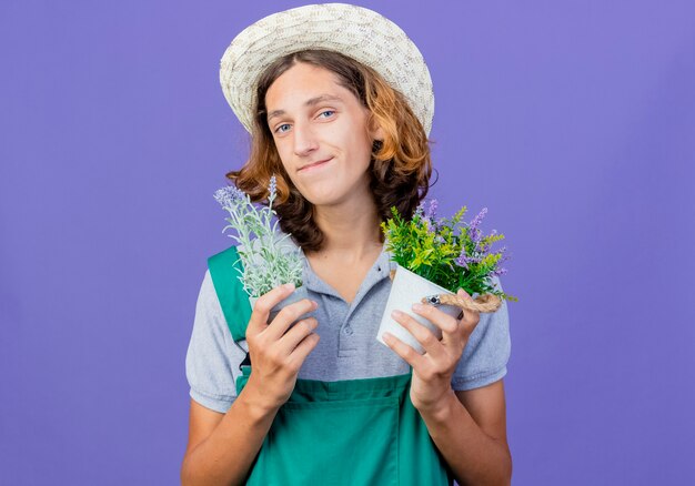 Hombre joven jardinero vestido con mono y sombrero sosteniendo plantas en macetas