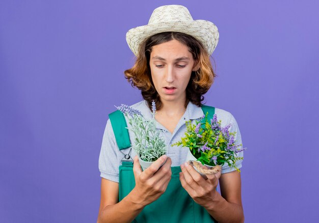 Hombre joven jardinero vestido con mono y sombrero sosteniendo plantas en macetas