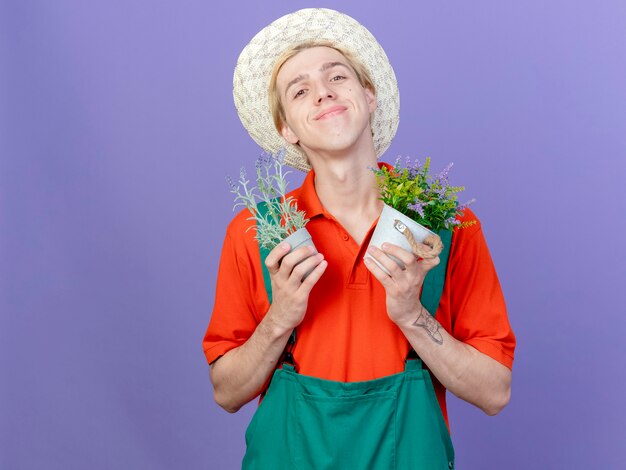 Hombre joven jardinero vestido con mono y sombrero sosteniendo plantas en macetas