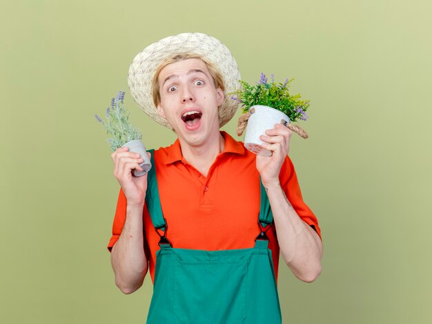 Hombre joven jardinero vestido con mono y sombrero sosteniendo plantas en macetas