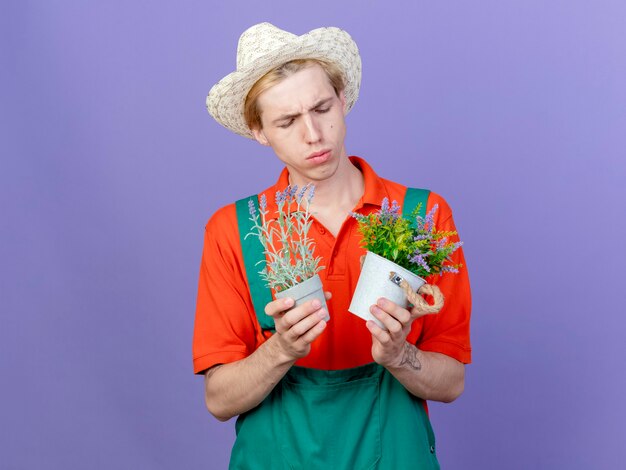 Hombre joven jardinero vestido con mono y sombrero sosteniendo plantas en macetas