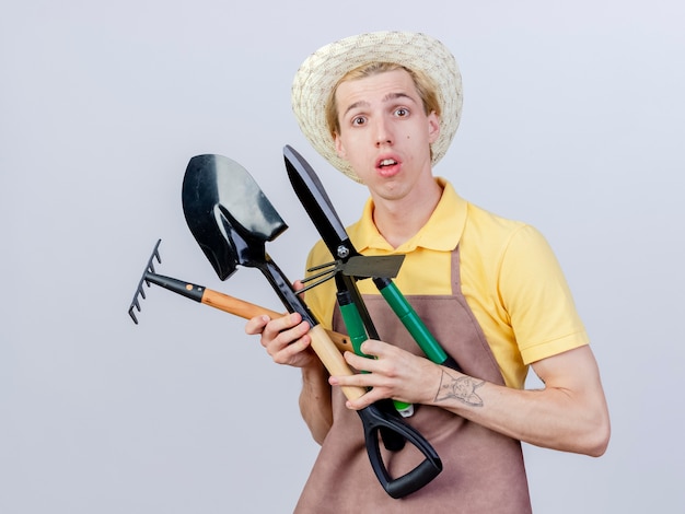 Foto gratuita hombre joven jardinero vestido con mono y sombrero sosteniendo rastrillo azadón y cortasetos sorprendido