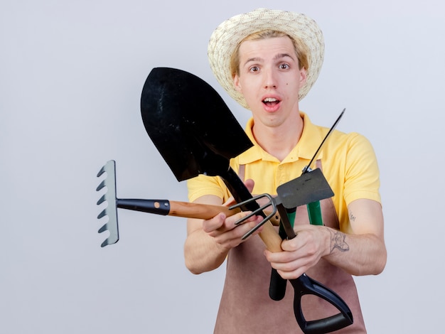 Foto gratuita hombre joven jardinero vestido con mono y sombrero sosteniendo rastrillo azadón y cortasetos sorprendido