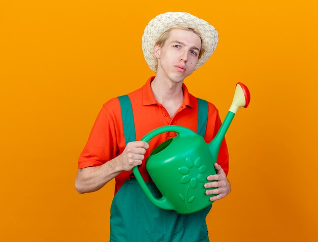 Hombre joven jardinero vestido con mono y sombrero sosteniendo regadera mirando a la cámara con cara seria de pie sobre fondo naranja