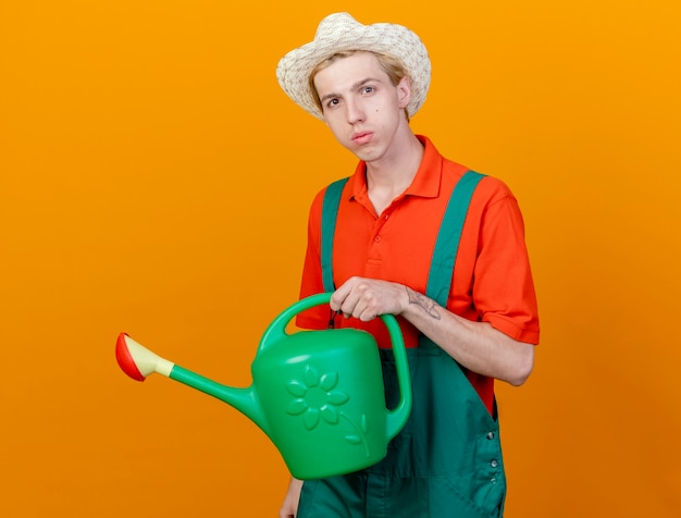 Hombre joven jardinero vestido con mono y sombrero sosteniendo regadera mirando a la cámara con cara seria de pie sobre fondo naranja