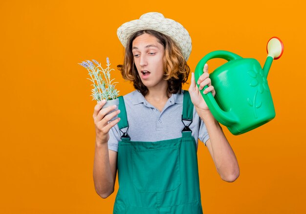 Hombre joven jardinero vestido con mono y sombrero sosteniendo regadera y planta en maceta