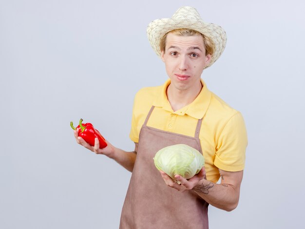 Hombre joven jardinero vestido con mono y sombrero sosteniendo repollo y pimiento rojo sonriendo