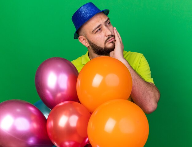 Hombre joven de lado mirando complacido con gorro de fiesta de pie detrás de globos poniendo la mano en la mejilla