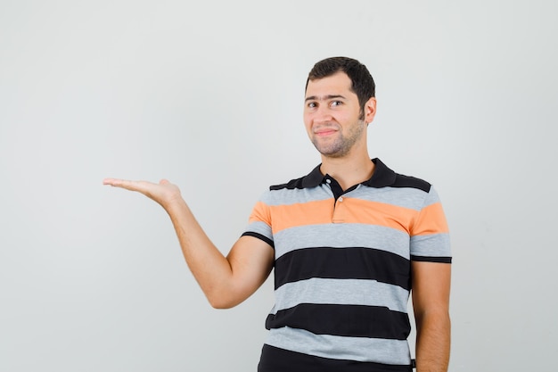 Hombre joven levantando la mano por mostrar algo en camiseta de rayas y luciendo satisfecho, vista frontal.