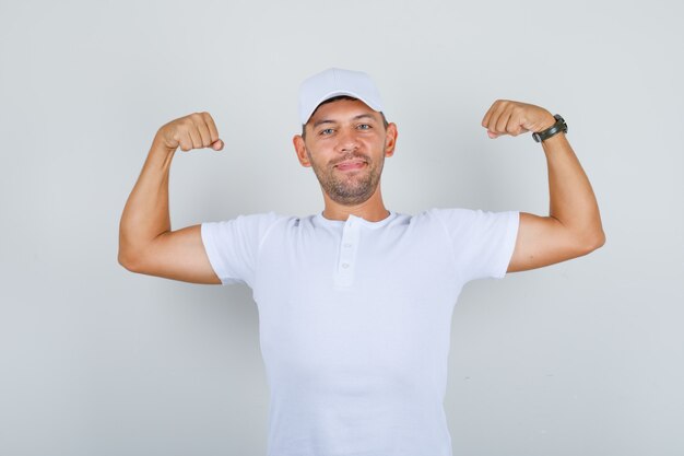 Hombre joven levantando las manos y mostrando los músculos en la camiseta blanca, gorra y mirando fuerte, vista frontal