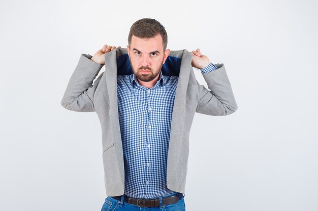 Foto gratuita hombre joven levantando las solapas de la chaqueta mientras posa en camisa, jeans, chaqueta de traje y mirando serio, vista frontal.