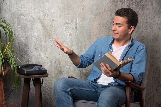 Hombre joven con libro sentado en una silla sobre fondo de mármol. Foto de alta calidad