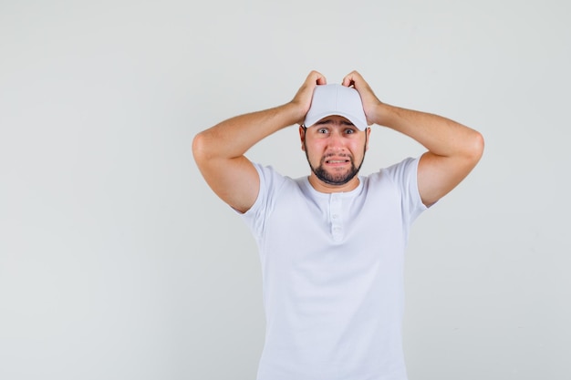 Hombre joven con la mano en la cabeza en camiseta blanca, gorra y mirando preocupado. vista frontal.