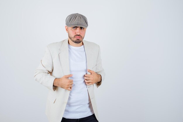 Hombre joven de la mano en la chaqueta con camiseta blanca, chaqueta y gorra gris y mirando serio. vista frontal.