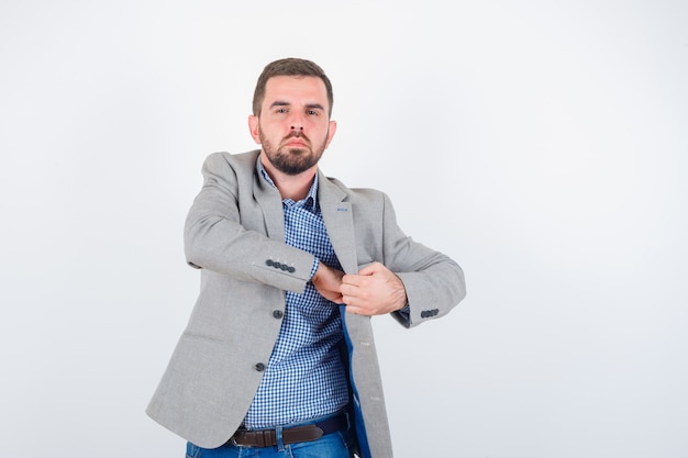 Hombre joven manteniendo la mano en el bolsillo de la chaqueta en camisa, jeans, chaqueta de traje y mirando confiado. vista frontal.