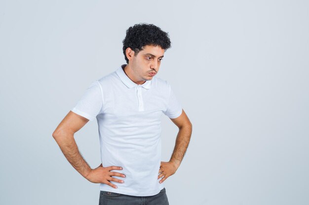 Hombre joven manteniendo las manos en la cintura en camiseta blanca, pantalones y mirando pensativo, vista frontal.
