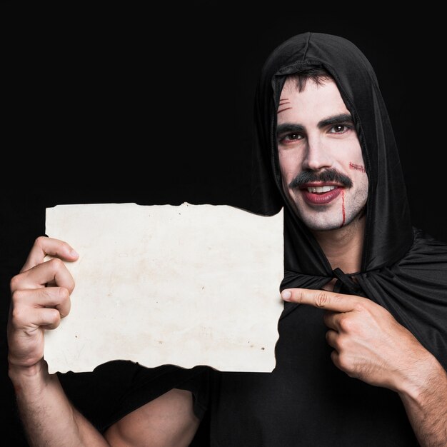 Hombre joven en manto negro y camiseta posando en estudio con un pedazo de papel en blanco