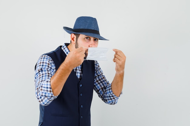 Hombre joven con máscara médica en camisa, chaleco, sombrero y mirando con cuidado.