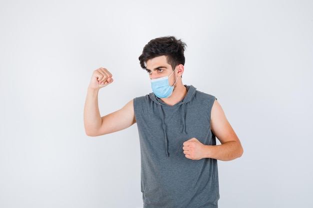 Hombre joven con máscara mientras muestra los músculos y aprieta el puño en una camiseta gris y se ve serio