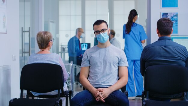 Hombre joven con mascarilla contra el coronavirus mirando a la cámara en la sala de espera del hospital. Mujer mayor con andador esperando consulta en la clínica.