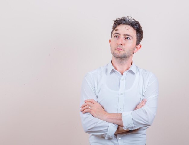 Hombre joven mirando hacia arriba con los brazos cruzados en camisa blanca y mirando pensativo