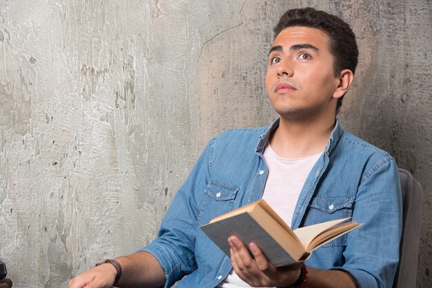 Hombre joven mirando hacia arriba con libro y sentado en una silla. Foto de alta calidad