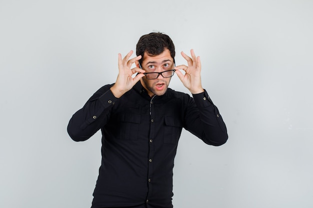 Hombre joven mirando por encima de las gafas con camisa negra y mirando curioso.