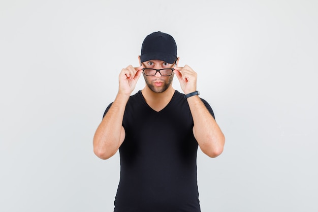 Hombre joven mirando por encima de las gafas en camiseta negra, gorra y mirando sorprendido.