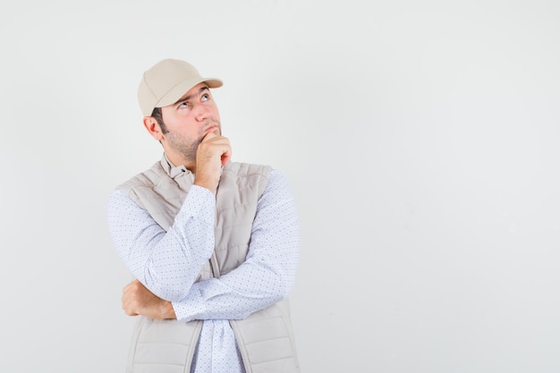 Hombre joven mirando a otro lado en camisa, chaqueta sin mangas, gorra y mirando pensativo, vista frontal. espacio para texto