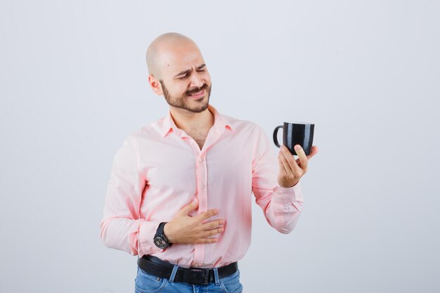 Hombre joven mirando la taza en camisa rosa, jeans y luciendo optimista. vista frontal.