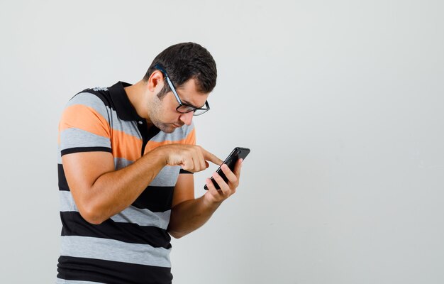 Hombre joven mirando el teléfono en camiseta, gafas y mirando con cuidado. espacio para texto