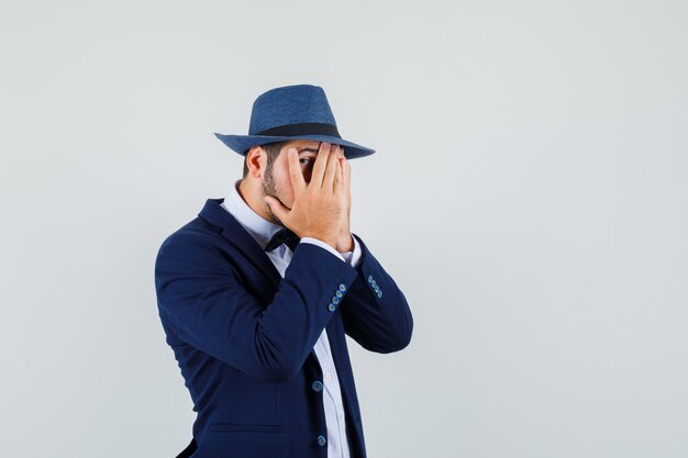 Hombre joven mirando a través de los dedos en traje, sombrero y mirando asustado, vista frontal.